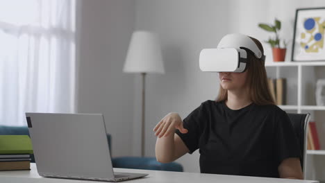 Junge-Benutzerin-Betrachtet-Den-Bildschirm-Eines-Laptops-Mit-Einem-Modernen-VR-Head-Mounted-Display-Und-Gestikuliert-Mit-Den-Händen,-Um-Das-Mittlere-Frauenporträt-Zu-Hause-Zu-Steuern