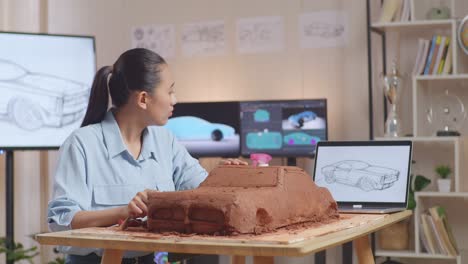 asian woman automotive designer looking at a laptop while working on the sculpture of car clay in the studio