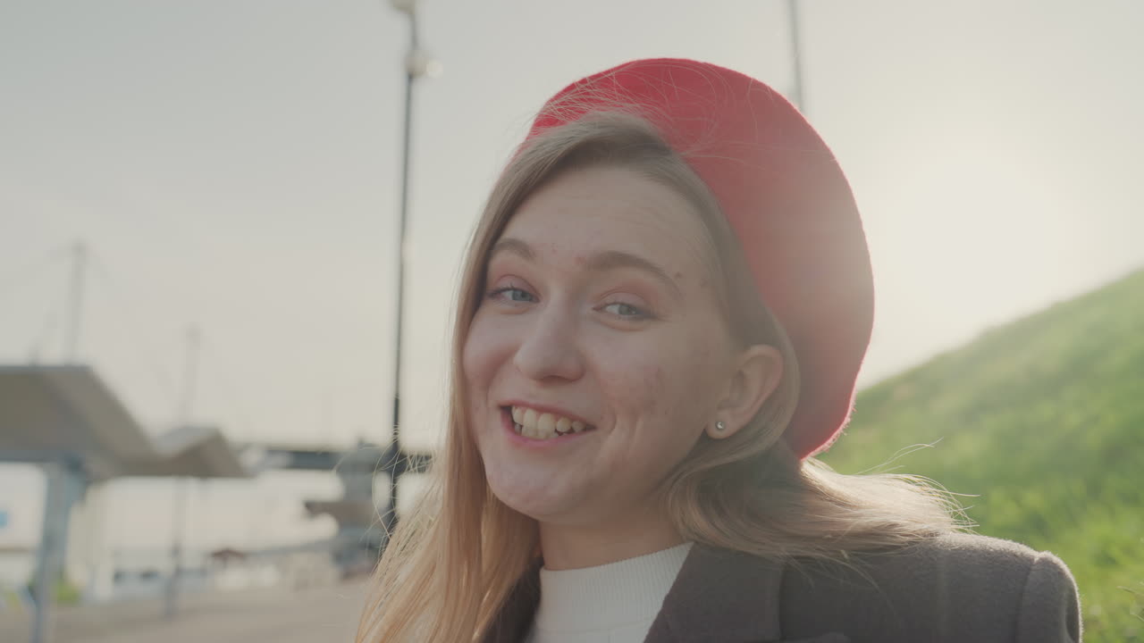 Ung kvinde iført rød baret smiler og sender et kys på en solbeskinnet vandpromenade, afslappet frakke, mild brise, hår i modlys, et oprigtigt portræt i vlog-stil der fanger glæde og legende charme, varmt gyldent lys.