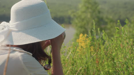 close up side view of woman wearing white hat and holding camera as she bends toward blooming yellow wildflower near hilltop with blurred river and forest background on calm summer day
