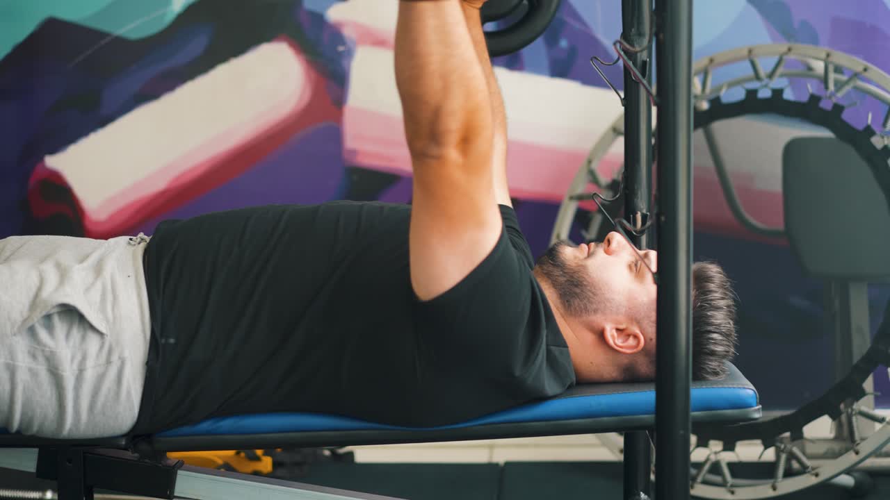 Arka planda trampolin bulunan bir spor salonunda, barbel ile bench press antrenmanı yapan genç bir adamın yandan görünüşü, hareketli çekim.