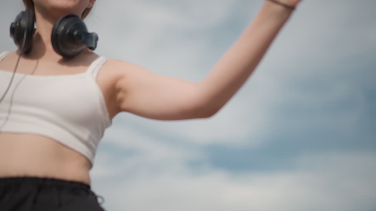 Bailarina aérea, mujer blanca, brazos abiertos, auriculares alrededor del cuello, top corto y pantalones holgados, movimiento fluido sobre un cielo azul nuboso, alcance expresivo y equilibrio elegante, pelo ondeando al viento, práctica en solitario