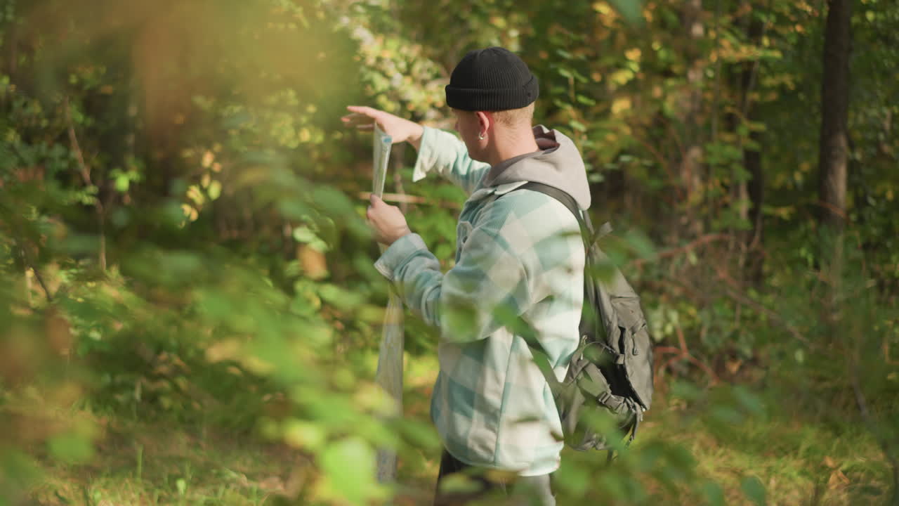 white hiker reading map in forest, backpacked navigator unfolds paper route between sunlit trees, scanning bearings and adjusting beanie while planning trail and checking foliage for landmarks, quiet