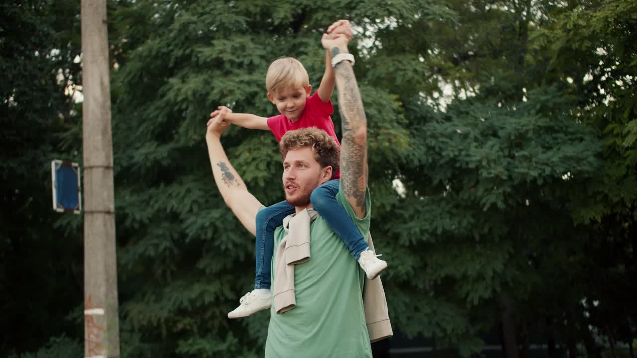 A happy father in a Green T-shirt and his blond son in a red T-shirt who sits on his dad s shoulders. Dad and son hold hands and spin with happiness in a green park
