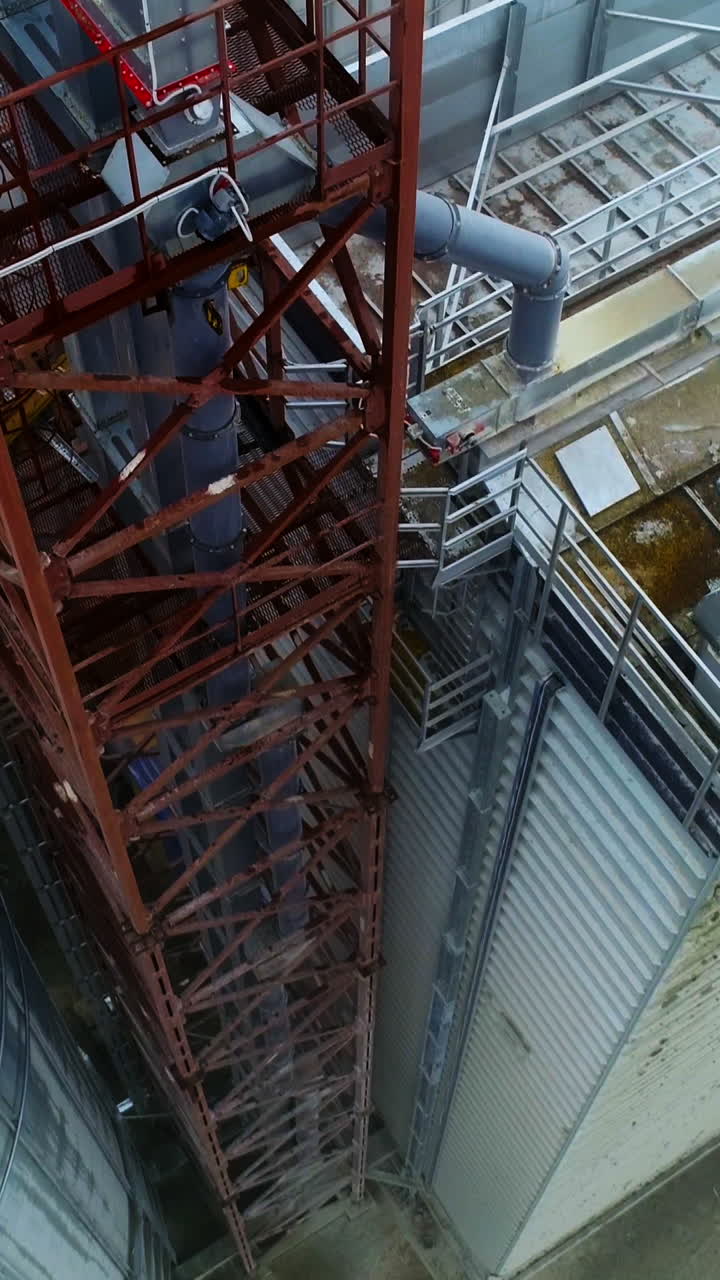 Metal supports near the huge elevator cisterns. Going down along the tower standing between the warehouse and grain tank. Aerial view. Vertical video