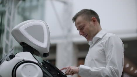 a man stands with a robot bot and asks him questions and asks for help by clicking on the screen on the robot body. human-robot interaction in the modern world