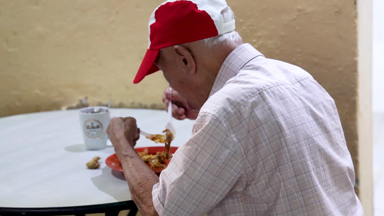 An elderly man in a red cap eats a meal at a round table in a simple, well-lit indoor setting, captured from a medium side angle