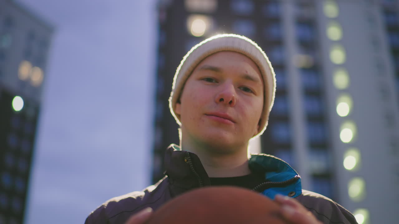 Giocatore urbano serale che tiene in mano un pallone da basket con controluce spettacolare, silhouette e bagliore sullo sfondo di torri residenziali, mani sul pallone e sguardo fisso sicuro, bokeh cinematografico e giacca texturizzata, a tarda ora