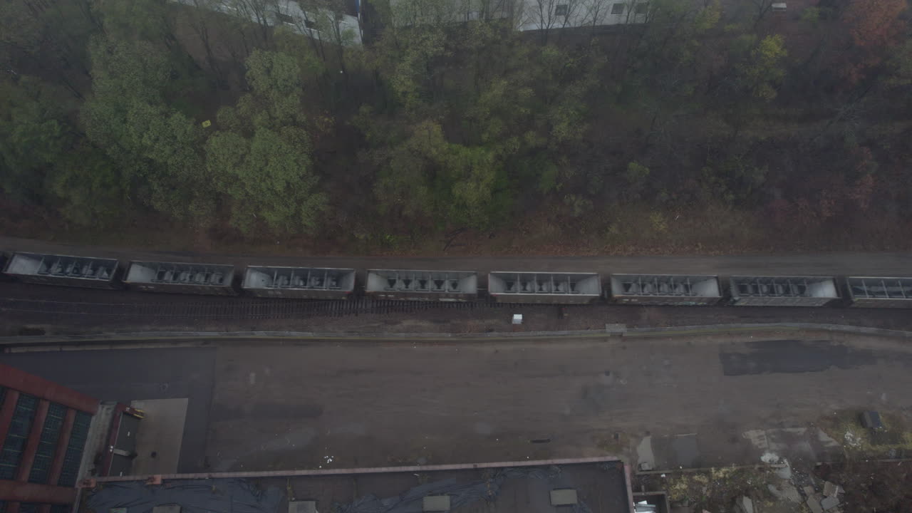 Aerial drone view of a freight train engine and cars passing through an industrial area bordered by trees and scattered debris