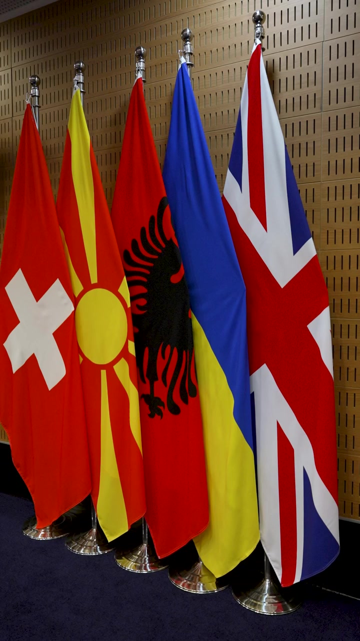 Flags of Switzerland, North Macedonia, Albania, Ukraine, and the United Kingdom are arranged in a conference room. These flags represent various nations and cultures.