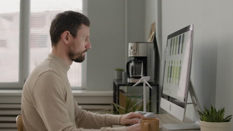 side view of man working with pc and drinking coffee sitting at desk in the office