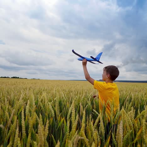 little boy plays on factory background. child in yellow t-shirt walking on wheat field and holds toy plane in his hand in summer.