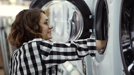 a young positive woman in a plaid shirt choosing washing machine in the shot of household appliances. look inside to the open