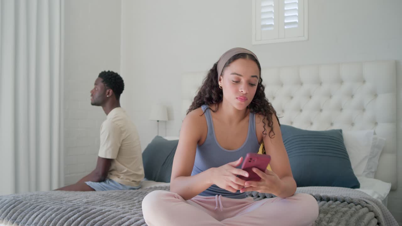 Coppia afroamericana seduta sul letto, la donna tiene in mano uno smartphone rosa e scorre, l'uomo rimane in disparte.
