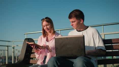 estudiantes de secundaria estudian al aire libre; hombre centrado en la computadora portátil, mujer mostrando algo en el libro contra el fondo con barandilla de hierro