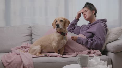 young sick woman checking her temperature with thermometer lying on sofa with her small dog, suffering from seasonal flu or cold. ill girl feel unhealthy with influenza at home