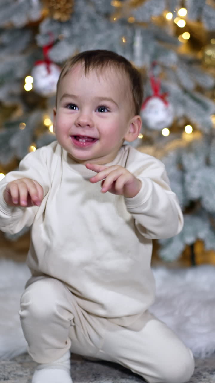 Smiling happy Caucasian child waving hands excitedly. Adorable baby boy sits on the floor near the Christmas tree. Vertical video