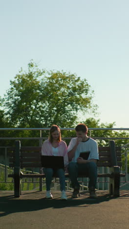 estudiantes de secundaria estudiando al aire libre, sentados en bancos, uno usando una computadora portátil y el otro sosteniendo una tableta, rodeados de barandillas, árboles y luz solar, creando una atmósfera de aprendizaje tranquila