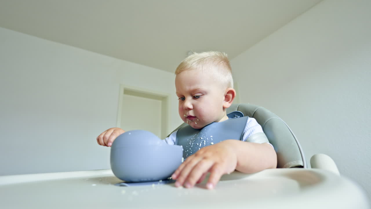 A little boy with a silicone apron sits in a high chair and eats porridge with a spoon on his own. Toddler eats breakfast on his own and chews happily. Safe silicone utensils for children