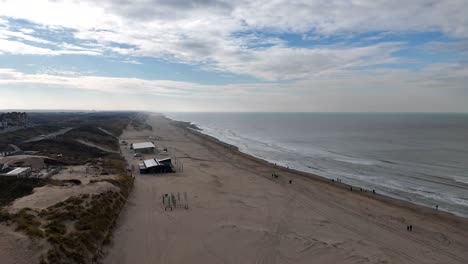 Aerial forward shot of sandy beach in Noordwjik with ocean and walking people on cloudy and sunny winter day. Durch Restaurants buildings beside dunes in Netherlands. Wide shot.