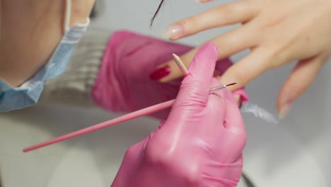 close up view of nail technician wearing pink glove using stick to remove excess red nail polish from side of client fingernail and gently cleaning stick against glove for precise detailing