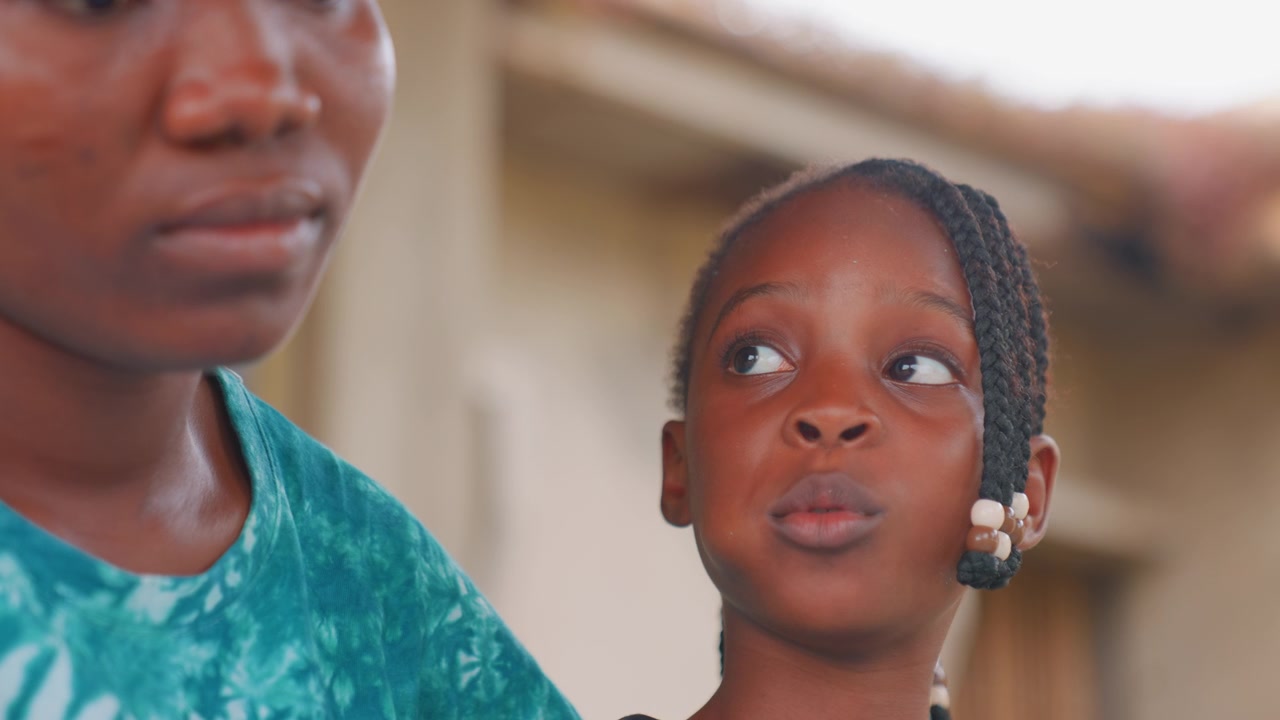Acercamiento de unión entre madre e hija afroamericanas. La madre observa a su hija con una mirada tierna mientras la niña examina un pequeño objeto, con su cabello trenzado. Luz natural cálida, un fondo residencial tranquilo y una crianza serena.