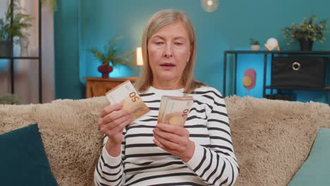 mature woman joyfully counting money cash on couch after lottery win happy with bonus gift at home