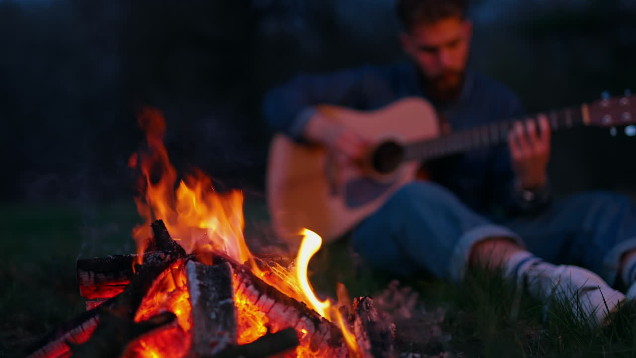Man sitting by bonfire with guitar. Lonely man sitting by bonfire on the beach near forest playing acoustic guitar