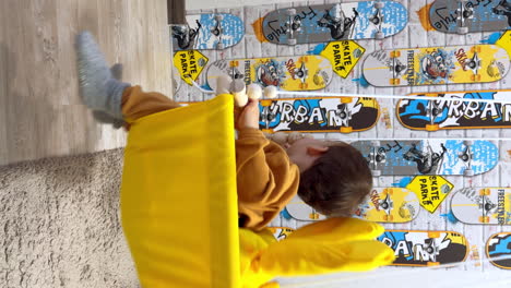 lovely child in yellow suit sits in the swing and waves his hand. sweet baby boy having fun time at home. vertical screen.
