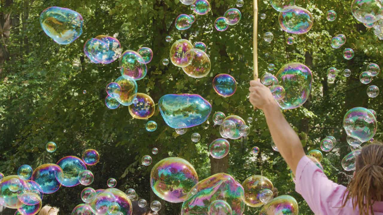 A person uses sticks and string to make giant, iridescent soap bubbles outdoors in a sunlit park, surrounded by green trees and natural light
