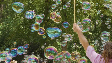 a person uses sticks and string to make giant, iridescent soap bubbles outdoors in a sunlit park, surrounded by green trees and natural light