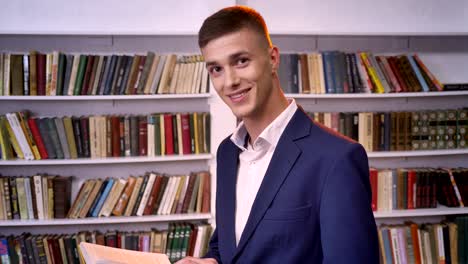 young handsome businessman is reading book, watching at camera, smiling, bookshelf on background, side view