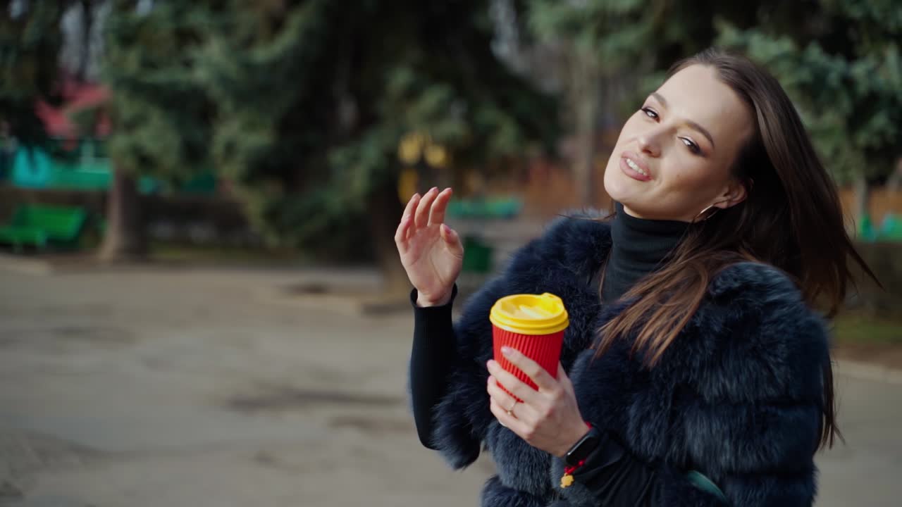 Fashionable girl in the park. Portrait of a lovely woman in fur coat with plastic cup of coffee on park background in sunny day. Slow motion.