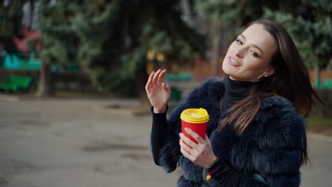 fashionable girl in the park. portrait of a lovely woman in fur coat with plastic cup of coffee on park background in sunny day. slow motion.