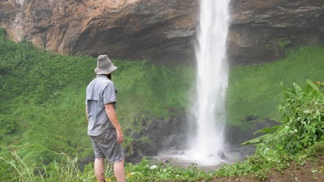 Un-Jengibre-Barbudo-Que-Lleva-Un-Sombrero-Bosquimano-Se-Encuentra-En-La-Base-De-Una-Poderosa-Caída-De-Agua-En-África-Tropical