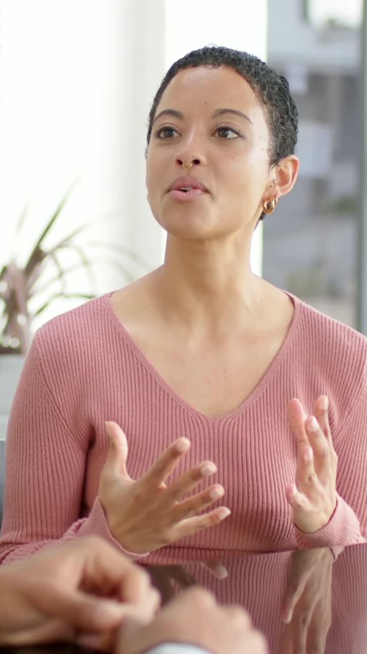 Vídeo vertical: Mãos surgindo, mulher fazendo uma observação à mesa com blusa rosa, pulseira visível.