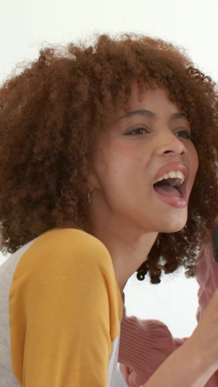 Vertical video: Singing woman in pink knit holding mic, friend with yellow sleeves joining at home