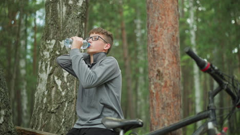 young boy wearing glasses and grey long-sleeve top drinks from a water bottle, taking a break in a forest surrounded by tall green trees, a bicycle is parked beside him in the background