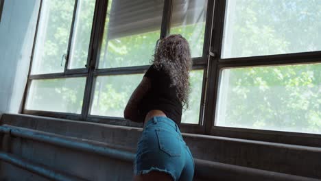 tattooed woman in hot pants posing confidently by industrial windows in natural light