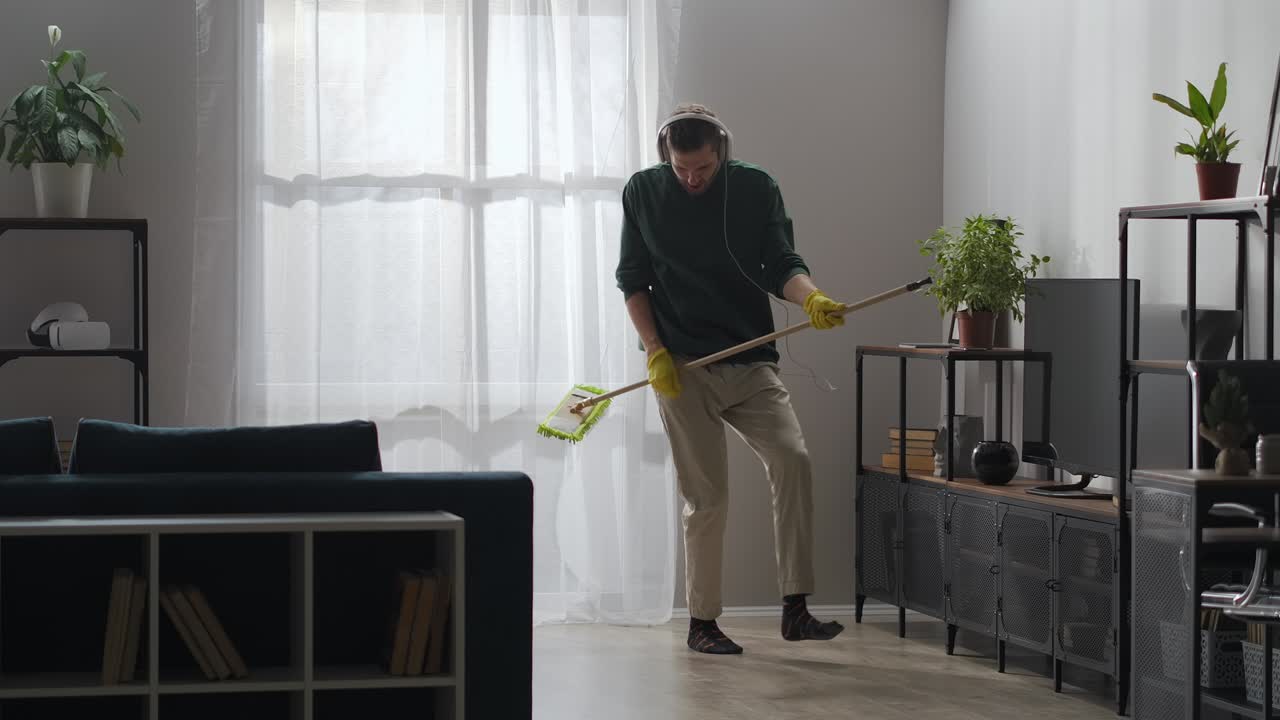 el hombre está haciendo la limpieza en casa y bailando con la mopa, lavando el suelo en la sala de estar, tocando la mopa como guitarra
