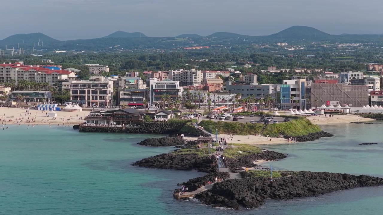 Inquadratura orizzontale con movimento a destra e panoramica a sinistra su Hamdeok Beach, Sinheung-ri, Isola di Jeju, Corea del Sud, che mostra acqua turchese, rocce vulcaniche, spiaggia ed edifici