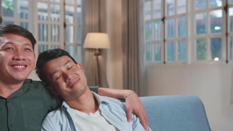 happy asian gay couple watching tv, while sitting on a couch in cozy stylish apartment. adult boyfriends hug, watching funny videos and streaming movies from online service