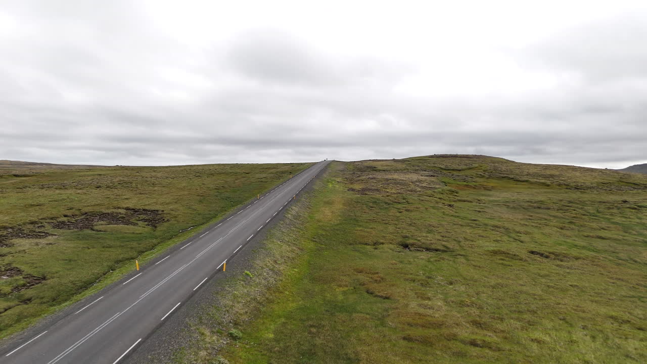 Dronebillede i fugleperspektiv af to cyklister, der kører langs en afsidesliggende kystvej i det østlige Island med fjorde, bjerge og åbent landskab under en overskyet himmel, hvilket symboliserer eventyrrejser, ensomhed og frihed.
