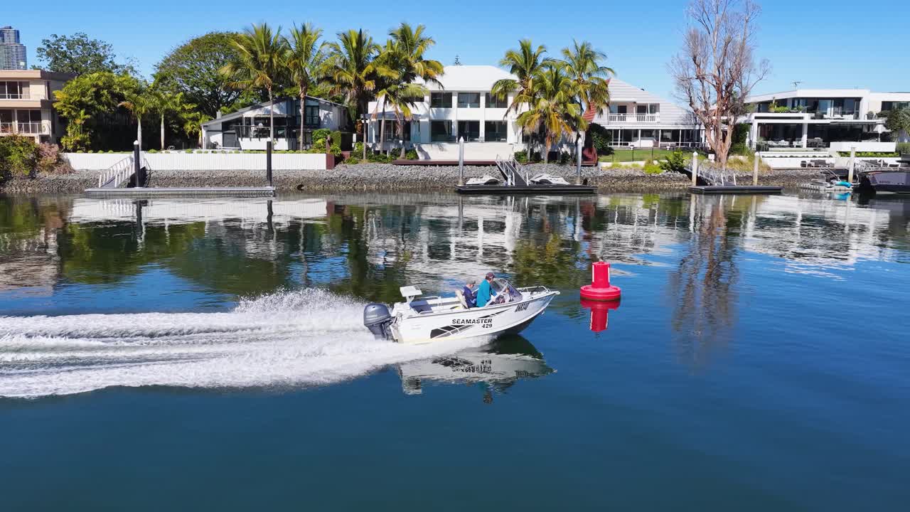 A small speedboat travels quickly along a calm canal bordered by modern waterfront houses under bright daylight, captured in smooth aerial drone footage