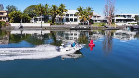 a small speedboat travels quickly along a calm canal bordered by modern waterfront houses under bright daylight, captured in smooth aerial drone footage