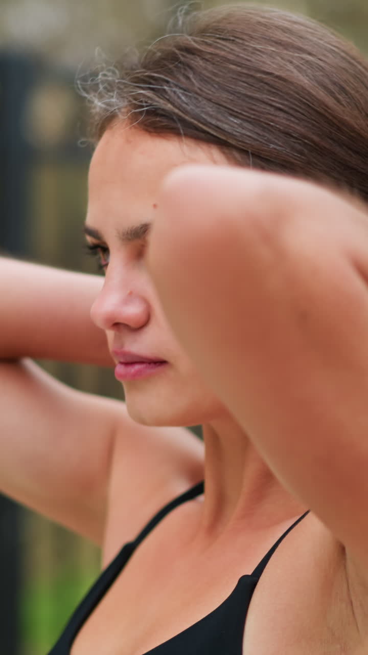 Close-up portrait of woman packing her hair while standing outdoors, woman tying back hair, preparing for exercise or workout, with a background of green foliage and iron fence