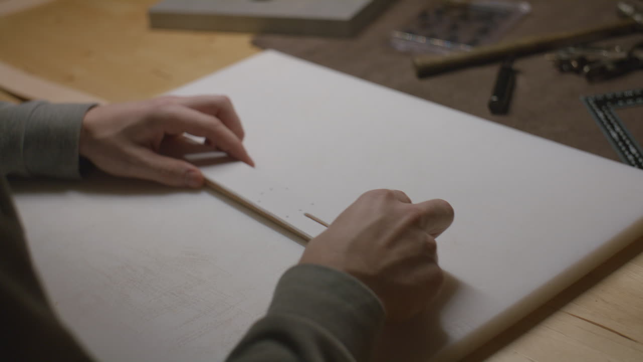 A close-up of hands working on a leather crafting project using templates and precision tools on a wooden workbench.