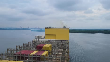 aerial footage shows a yellow-funnel container vessel departing contecon terminal, gliding down ecuador’s guayas river past guayaquil under brooding clouds