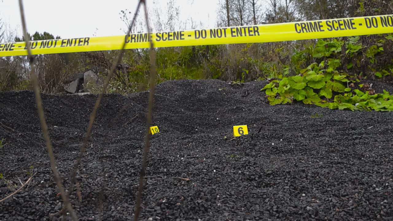 Yellow police line or forensic investigation crime scene tape pulled in front of bullets and other evidence next to crime numbered markers on dark gravel ground outdoors in nature, bokeh background