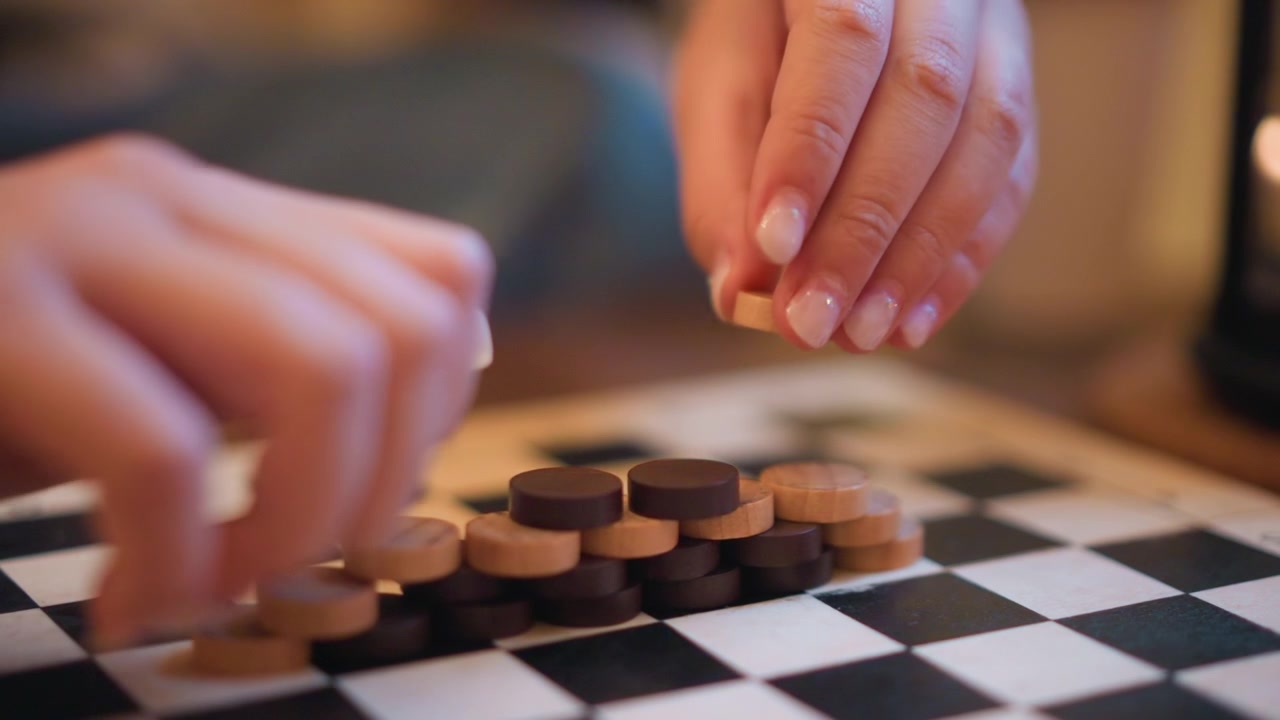 Nahaufnahme von Händen, die hölzerne Dame-Steine auf einem Schachbrett stapeln und bewegen. Der Fokus liegt ausschließlich auf der taktilen Interaktion zwischen Fingern und Spielsteinen aus Holz in hellbraunen und dunklen Farben.